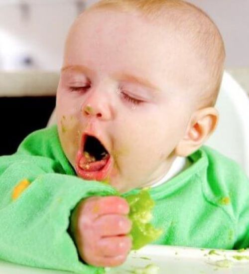 A baby sitting, coughing in a high chair eating food