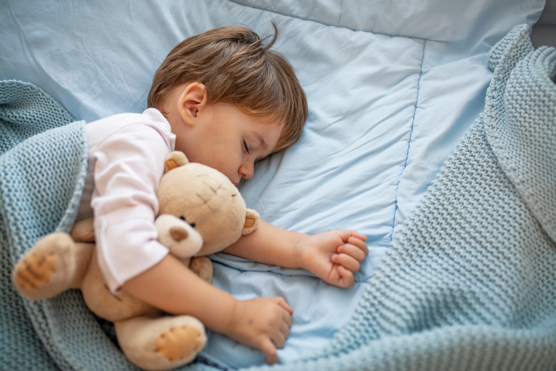 Little boy sleeping in bed with a teddy bear on blue bedding